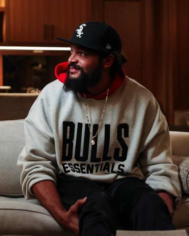 . A man in a Bulls Essentials sweatshirt sits comfortably on a couch, relaxed and casual in his home environment.