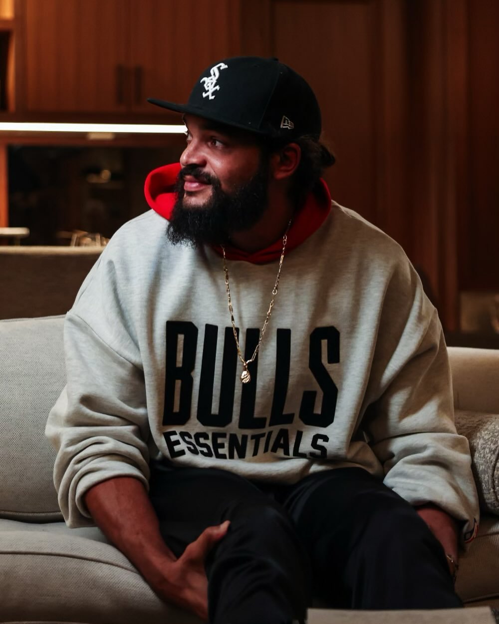 . A man in a Bulls Essentials sweatshirt sits comfortably on a couch, relaxed and casual in his home environment.